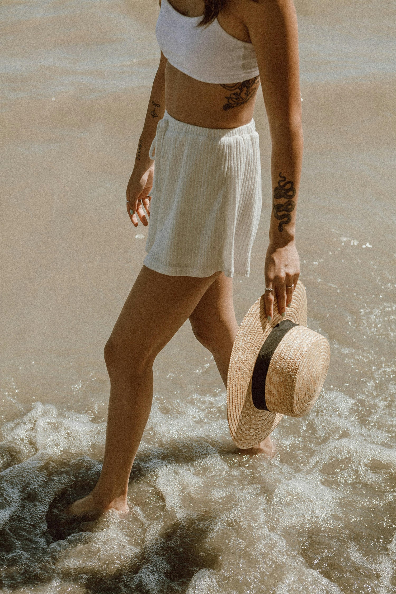Woman walking along the shoreline carrying a sun hat